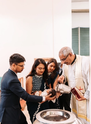 Photograph of a baby baptism ceremony in Pune, India, showing a priest blessing a small baby held by a parent, surrounded by family members in a traditional setting.