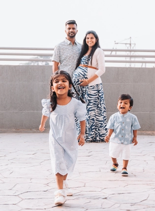 Image from a maternity photoshoot featuring a pregnant mother and father standing together and smiling in the background, while their two young children run joyfully toward the camera, captured in Pune, India.