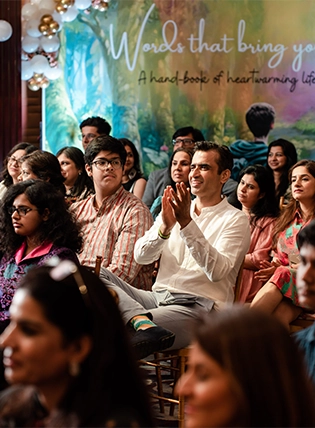 Photograph taken during a corporate event in Pune, India, showing a person applauding enthusiastically among a group of other attendees in the background. The audience is seated, and the atmosphere suggests a positive and engaging event