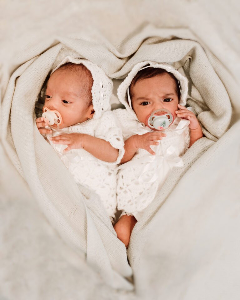 Candid picture of twin newborn children covered in a white cloth shaped like a heart.